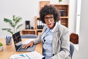 Poster - black woman with curly hair wearing call center agent headset at the office in shock face, looking s