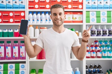 Canvas Print - Young caucasian man working at pharmacy drugstore showing smartphone screen smiling happy pointing with hand and finger to the side