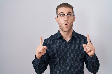 Canvas Print - Young caucasian man standing over isolated background amazed and surprised looking up and pointing with fingers and raised arms.