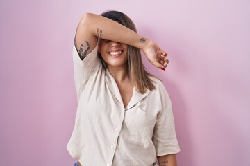 Poster - Blonde woman standing over pink background covering eyes with arm smiling cheerful and funny. blind concept.