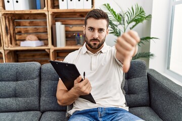 Canvas Print - Handsome hispanic man holding clipboard working at psychology clinic looking unhappy and angry showing rejection and negative with thumbs down gesture. bad expression.