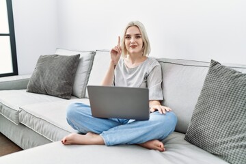 Poster - Young caucasian woman using laptop at home sitting on the sofa showing and pointing up with finger number one while smiling confident and happy.