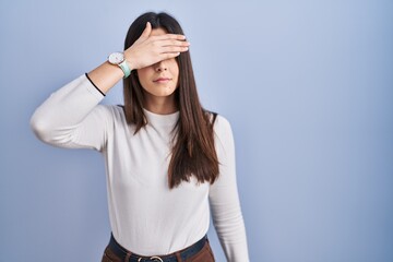 Sticker - Young brunette woman standing over blue background covering eyes with hand, looking serious and sad. sightless, hiding and rejection concept