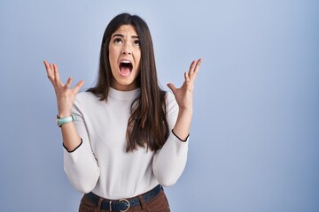 Poster - Young brunette woman standing over blue background crazy and mad shouting and yelling with aggressive expression and arms raised. frustration concept.