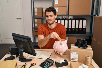 Canvas Print - Young hispanic man working at small business ecommerce holding piggy bank clueless and confused expression. doubt concept.