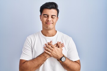 Poster - Hispanic man standing over blue background smiling with hands on chest with closed eyes and grateful gesture on face. health concept.