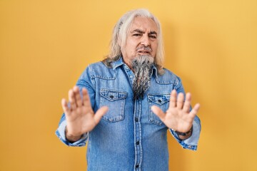 Middle age man with grey hair standing over yellow background moving away hands palms showing refusal and denial with afraid and disgusting expression. stop and forbidden.