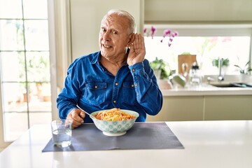 Canvas Print - Senior man with grey hair eating pasta spaghetti at home smiling with hand over ear listening an hearing to rumor or gossip. deafness concept.