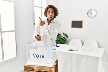 Poster - Young hispanic man voting putting envelop in ballot box surprised pointing with finger to the side, open mouth amazed expression.