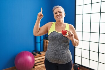 Sticker - Middle age woman wearing sportswear holding red heart smiling with an idea or question pointing finger with happy face, number one