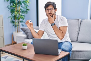 Poster - Handsome latin man holding virtual currency bitcoin using laptop serious face thinking about question with hand on chin, thoughtful about confusing idea