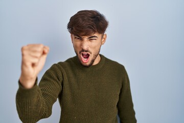 Sticker - Arab man with beard standing over blue background angry and mad raising fist frustrated and furious while shouting with anger. rage and aggressive concept.