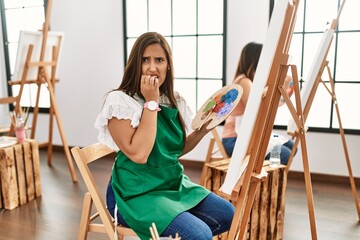 Sticker - Young hispanic artist women painting on canvas at art studio looking stressed and nervous with hands on mouth biting nails. anxiety problem.