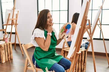 Poster - Young hispanic artist women painting on canvas at art studio with hand on chin thinking about question, pensive expression. smiling and thoughtful face. doubt concept.