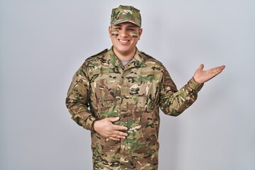 Canvas Print - Hispanic young man wearing camouflage army uniform smiling cheerful presenting and pointing with palm of hand looking at the camera.