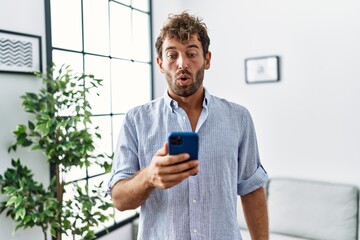 Sticker - Young handsome man using smartphone at home scared and amazed with open mouth for surprise, disbelief face