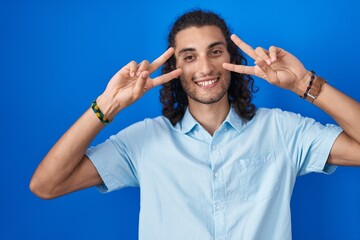 Poster - Young hispanic man standing over blue background doing peace symbol with fingers over face, smiling cheerful showing victory