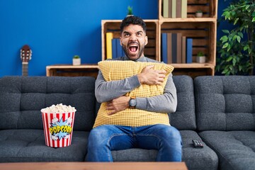 Wall Mural - Hispanic man with beard eating popcorn watching a movie at home angry and mad screaming frustrated and furious, shouting with anger looking up.