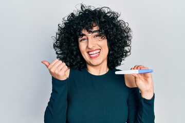 Poster - Young middle east woman holding pregnancy test result pointing thumb up to the side smiling happy with open mouth