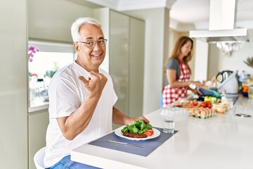 Wall Mural - Middle age hispanic couple eating healthy meal at home pointing thumb up to the side smiling happy with open mouth