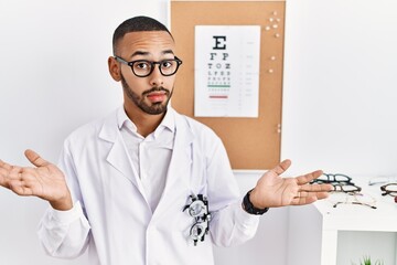 Poster - African american optician man standing by eyesight test clueless and confused expression with arms and hands raised. doubt concept.