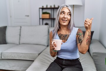 Canvas Print - Middle age grey-haired woman drinking coffee sitting on the sofa at home smiling with happy face looking and pointing to the side with thumb up.