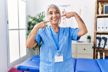 Poster - Middle age grey-haired woman wearing physiotherapist uniform at medical clinic smiling cheerful showing and pointing with fingers teeth and mouth. dental health concept.