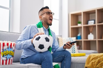 Poster - Young hispanic man football hooligan holding ball supporting team angry and mad screaming frustrated and furious, shouting with anger looking up.