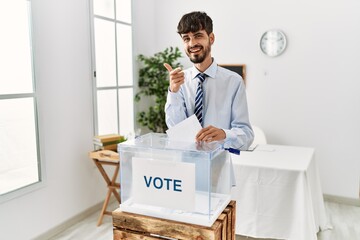 Poster - Hispanic man with beard voting putting envelop in ballot box pointing fingers to camera with happy and funny face. good energy and vibes.