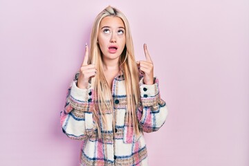 Wall Mural - Young blonde girl wearing casual clothes amazed and surprised looking up and pointing with fingers and raised arms.