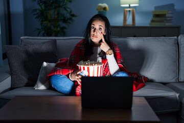 Sticker - Hispanic woman eating popcorn watching a movie on the sofa pointing to the eye watching you gesture, suspicious expression