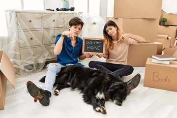 Poster - Young caucasian couple with dog holding our first home blackboard at new house pointing down looking sad and upset, indicating direction with fingers, unhappy and depressed.