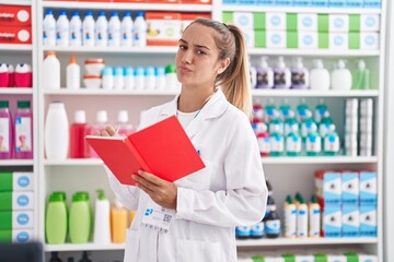 Wall Mural - Young blonde woman working at pharmacy drugstore holding notebook smiling looking to the side and staring away thinking.