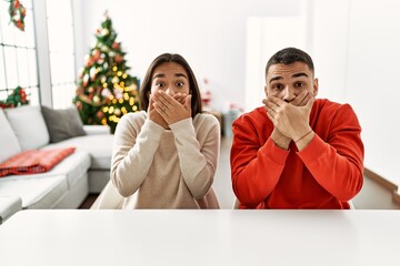 Sticker - Young hispanic couple sitting on the table by christmas tree shocked covering mouth with hands for mistake. secret concept.