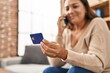 © Krakenimages.com - Middle age hispanic woman talking on the smartphone and using credit card sitting on sofa at home