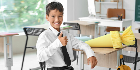 Wall Mural - Happy little boy with school cone showing thumb-up gesture in classroom