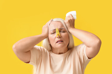 Poster - Ill mature woman with clothespin and tissue on yellow background. Stuffy nose concept