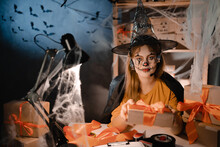 Startup Small Business Entrepreneur Or Freelance Caucasian Woman In Halloween Makeup Sitting At Table Among Boxes Holding Out One With Orange Bow. Online Marketing Packaging Box And Shipping