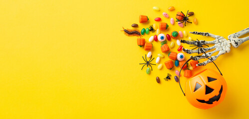 Wall Mural - Halloween decorations concept. Top view photo of skeleton hand holding pumpkin basket for trick-or-treat with candies eyes insects centipede and spiders on isolated yellow background