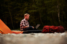 Businessman Using Laptop Sitting On Inflatable Mattress At Campsite