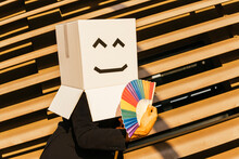 Businessman Wearing Box With Smiley Face Holding Rainbow Color Hand Fan On Sunny Day