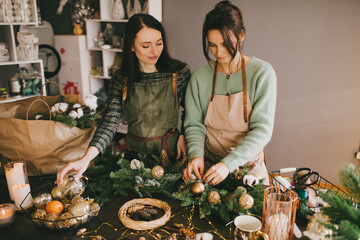 Wall Mural - Two millennial women making Christmas wreath using pine branches and festive decorations. Small business