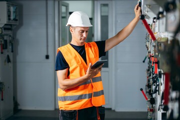 Adult electrician builder engineer testing and screwing equipment in fuse box and repairing of modern electricity power station using data from the tablet. Automatic control cabinet