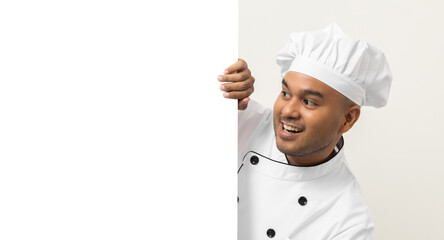 Wall Mural - Happy Young handsome asian man chef in uniform looks out from behind an empty whiteboard. Cooking indian man hiding behind big blank billboard for advertising text menu in kitchen and restaurant.