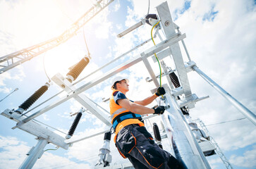 Electrician in protective helmet working on high voltage power lines. Highly skilled workmen servicing the electricity grid. Modern power station with power towers