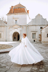 A beautiful red-haired bride in a satin white dress with a veil poses in the courtyard of an ancient castle.