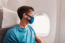 Young Man Travels By Airplane Flight, Wears A Face Mask During The Covid-19 Pandemic And Listens To Music With Headphones And Looks Out The Window