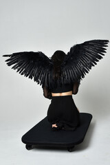 Wall Mural - full length portrait of beautiful asian model with dark hair, wearing black gothic skirt costume, angel feather wings with horned headdress. kneeling  pose  isolated on studio background.
