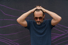 Thoughtful Mature Man Standing With Hand Folded Above His Head Next To Dark Urban Wall Leaned Head Down Wearing Sunglasses, Dark Blue T-shirt And Light Shorts. Stress, Middle Age Crisis Man Concept