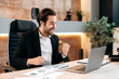 © Kateryna - Big profit, success. Happy cheerful caucasian businessman, office worker, sits at a workplace in office, works in a laptop, gesticulates with his hands, rejoices in a big profit, a good deal, smiling